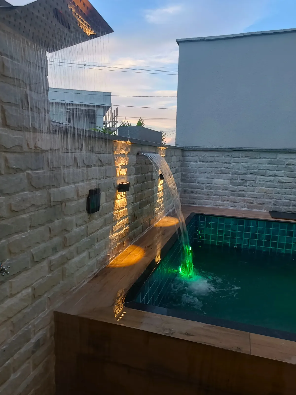 Piscina com cascata iluminada e parede de pedra, ao entardecer